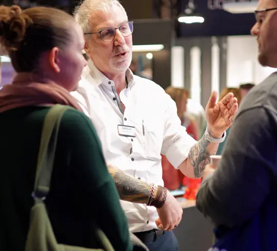 Ein Mann mit kurzen grauen Haaren und Bart unterhält sich mit einer Frau und einem Mann auf einer Messe