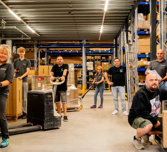 Eine Gruppe von Mitarbeitern in einem Lager, umgeben von Regalen mit Kartons. Zwei Personen bedienen Palettenwagen, während andere sich an eine Leiter lehnen oder im Vordergrund sitzen. Alle wirken beschäftigt, aber gut gelaunt.