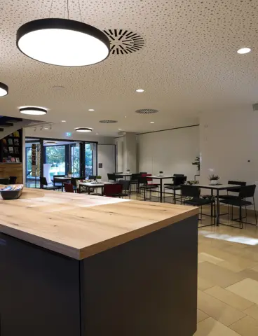 Das Bild zeigt einen modern gestalteten Innenraum mit runden Deckenleuchten, die den Raum gleichmäßig ausleuchten. Im Vordergrund steht eine Kücheninsel mit Holzarbeitsplatte, während im Hintergrund ein Sitzbereich mit Tischen und Stühlen sichtbar ist. Die Einrichtung wirkt einladend und funktional.