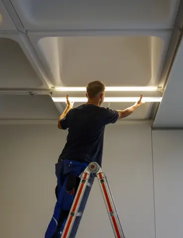 Ein Arbeiter auf einer Leiter installiert eine QUICK-FIXdc Deckenlampe in einem Raum mit moderner Rasterdecke.