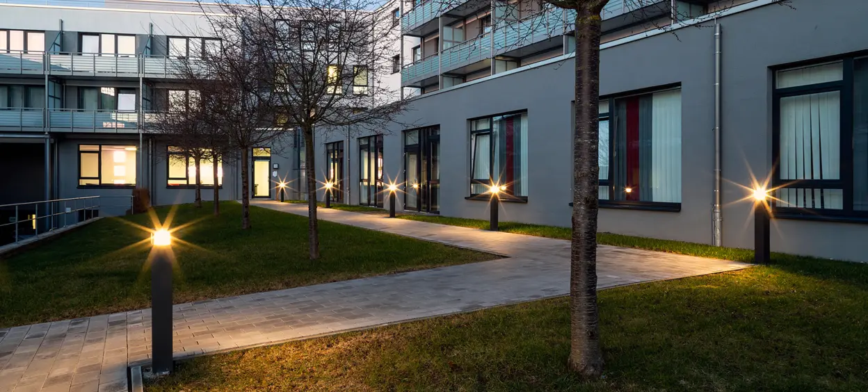 Abendaufnahme eines begrünten Innenhofs mit beleuchteten Wegleuchten und einem modernen Gebäudekomplex im Hintergrund.