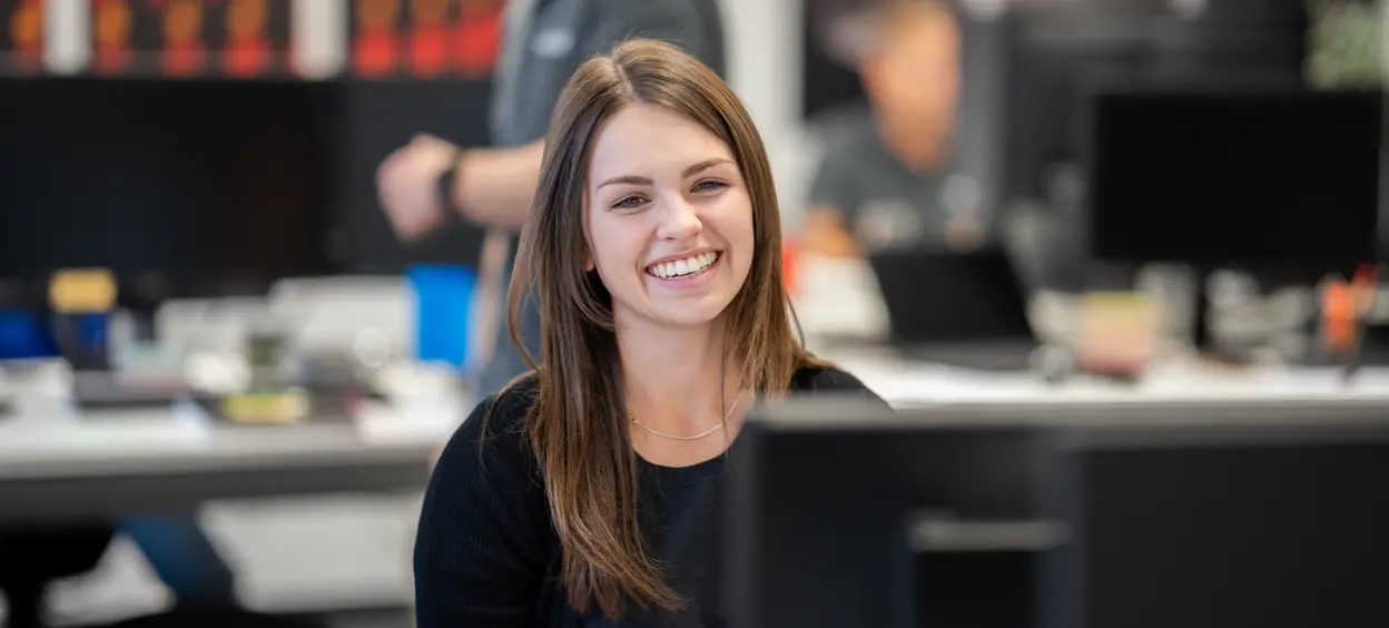 Eine junge Frau mit langen braunen Haaren lächelt herzlich in einem modernen Büro, während unscharf Kollegen im Hintergrund zu sehen sind.