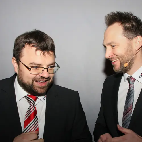 Ein Mann und eine Frau lachen herzlich, während sie sich in einer modernen Küche an einer Kaffeemaschine befinden.