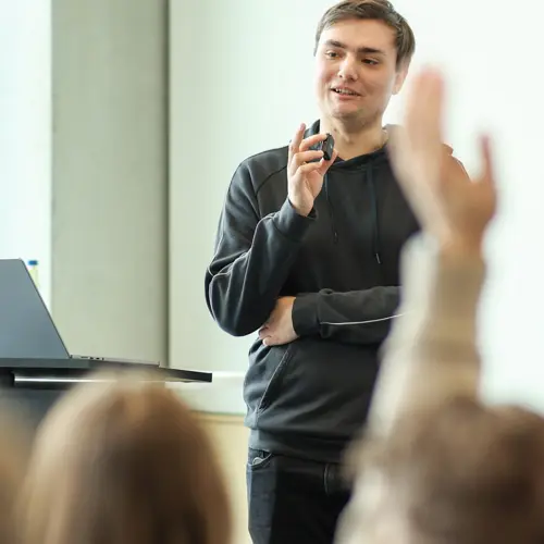 A woman in DOTLUX clothing holds a notebook and speaks to colleagues in a meeting room, who listen attentively.