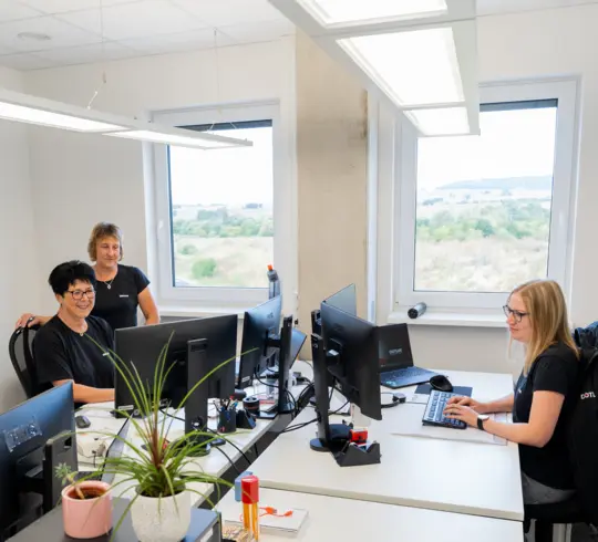 Drei lächelnde Frauen in einheitlichen schwarzen Shirts in einem modernen Büro.