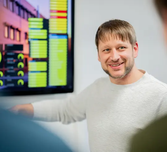 Ein Mann mit blonden Haaren und Bart erklärt an einem Monitor Grafiken. Zwei Personen stehen unscharf im Vordergrund