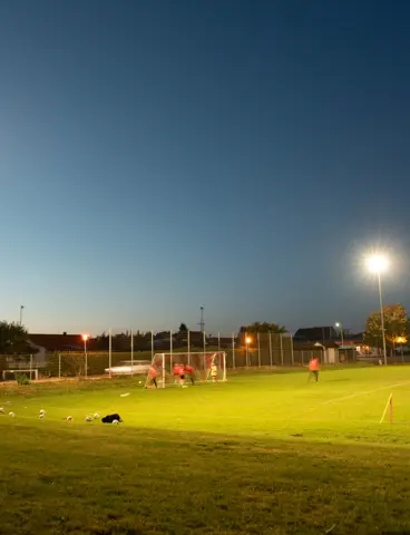 Ein beleuchteter Fußballplatz bei Dämmerung. Flutlichtmasten werfen helles Licht auf das Spielfeld, während Spieler das Tortraining durchführen. Im Hintergrund sind Bäume, Zäune und Gebäude zu erkennen.