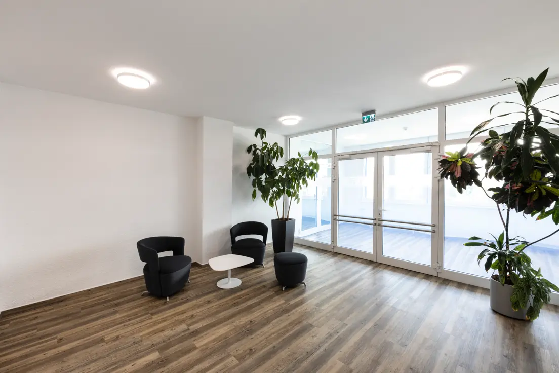 The entrance to an administrative building, featuring a seating area, plants, a glass façade and three ceiling lights