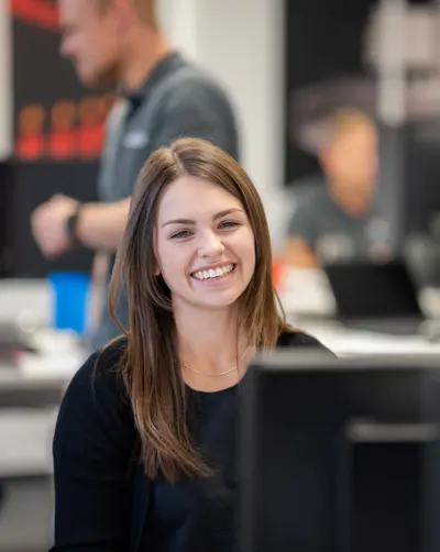 Eine junge Frau mit langen braunen Haaren lächelt herzlich in einem modernen Büro, während unscharf Kollegen im Hintergrund zu sehen sind.