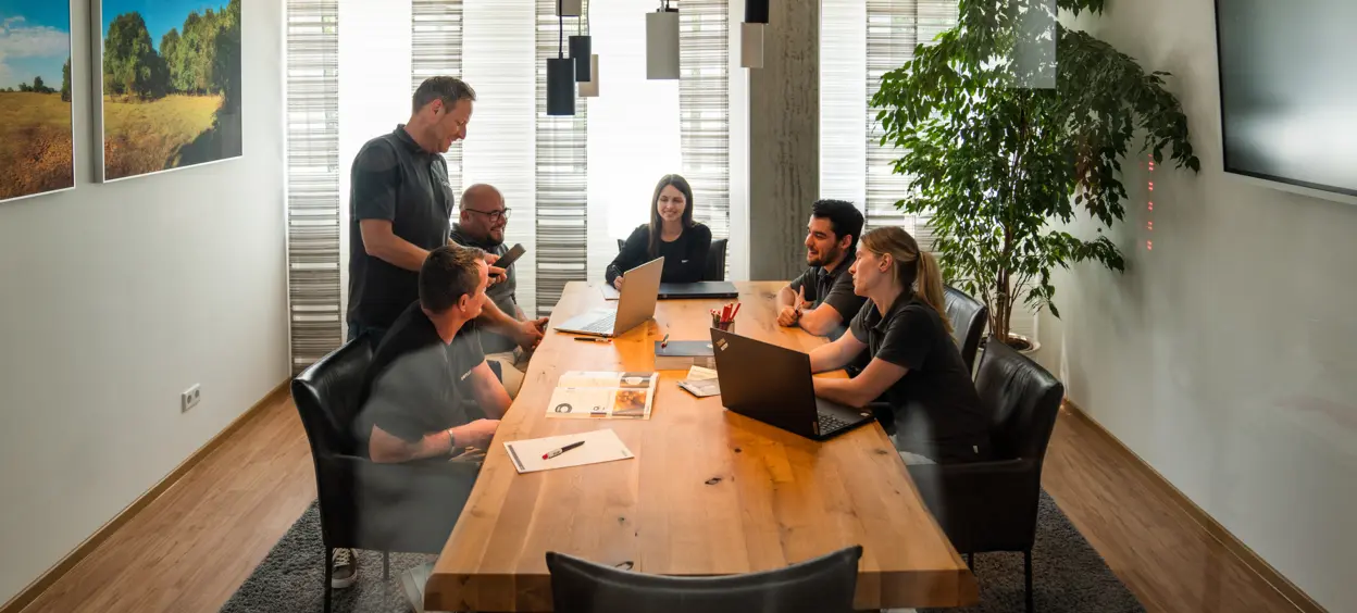 Gruppe von sieben Personen in einem modernen Besprechungsraum, teils mit Laptops arbeitend, umgeben von Pflanzen und Landschaftsbildern.