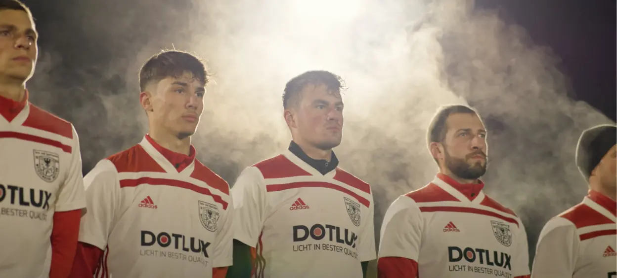 Vier Fußballspieler in weißen Trikots mit roten Akzenten und dem Schriftzug "DOTLUX" stehen unter hellem Flutlicht, das in einer kalten, dampfenden Atmosphäre leuchtet.