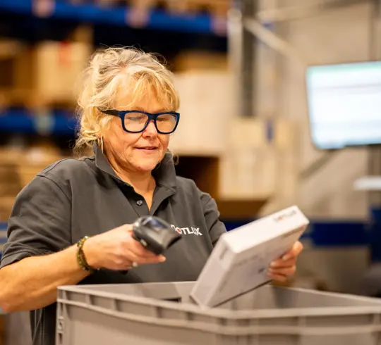 Eine Frau mit langen blonden Haaren scannt eine Verpackung. Sie steht in einer Lagerhalle