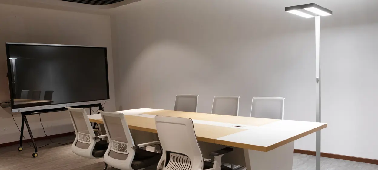 Ein moderner Konferenzraum mit großem Tisch, weißen Bürostühlen, ASSISTsensor-Stehleuchte und schwarzem Monitor auf einem Gestell.