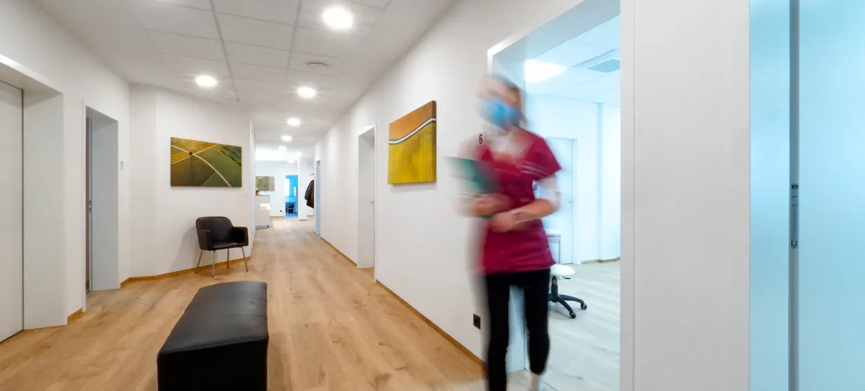 Ein heller, moderner Flur in einer Praxis oder Klinik, mit Holzfußboden, minimalistischen Möbeln und beleuchteten Kunstwerken an den Wänden.