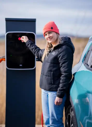 Eine Frau mit orangefarbener Mütze und schwarzer Jacke steht neben einem Elektroauto an einer Ladestation im Freien. Sie hält einen Stecker oder ein Kabel in der Hand, um das Fahrzeug aufzuladen.