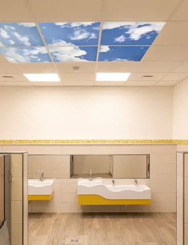 Ein modernes Badezimmer mit gelb-weißen Fliesen, Holzoptik-Türen und einer Deckenbeleuchtung, die blauen Himmel mit Wolken simuliert.