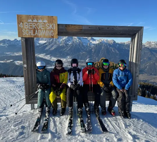 Sechs Skifahrer, die in einem großen Bilderrahmen auf der Skipiste vor einem Bergpanorama in Schladming sitzen