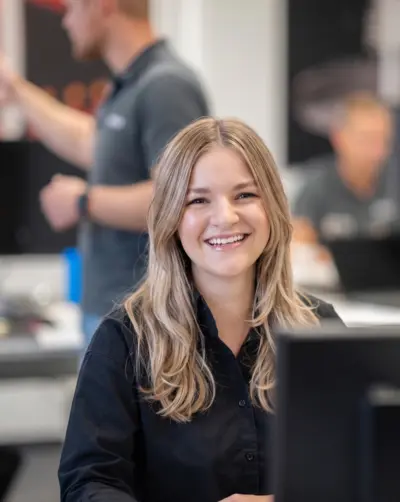 Une jeune femme aux longs cheveux bruns sourit chaleureusement dans un bureau moderne, tandis que des collègues apparaissent flous à l'arrière-plan.