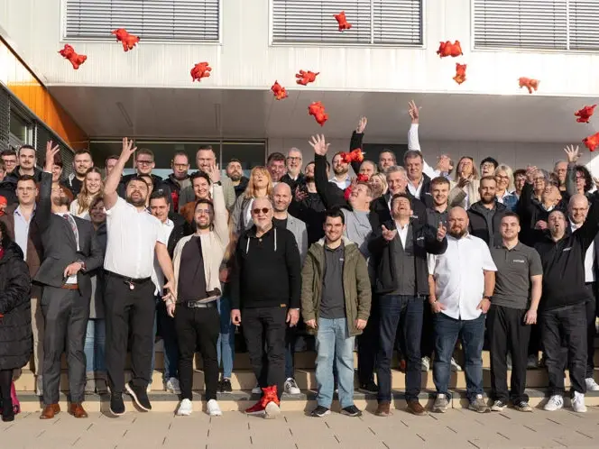 Gruppenfoto einer fröhlichen Menschenmenge vor einem Gebäude, die rote Stofftiere in die Luft wirft