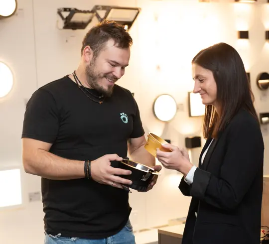 Ein Mann mit kurzen dunklen Haaren und eine Frau mit langen dunklen Haaren besprechen die CIRCLEvario. Der Mann hält die Leuchte in der Hand, die Frau eine goldfarbene, passende Blende.