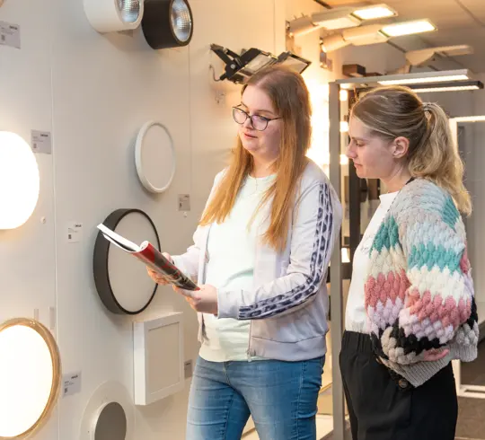 Zwei Frauen besprechen Leuchten an einer Wand. Eine hält einen Katalog in der Hand.